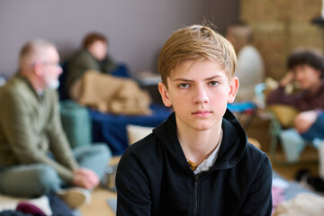 Serious schoolboy looking at camera while sitting against other refugees and temporarily homeless...