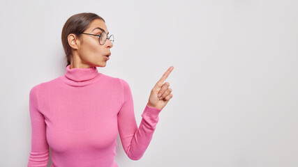 Studio shot of amazed brunette woman has wondered expression wears round spectacles and pink turtleneck points index finger on copy space demonstrates amazing offer isolated over white studio wall