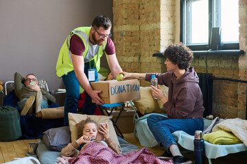 Young male volunteer with donation box bending in front of female refugee taking fresh green apple...