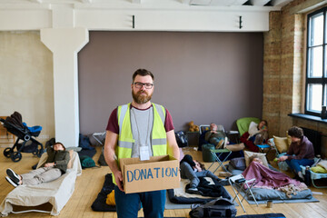 Male volunteer with donation box looking at camera against temporarily homeless people resting on sleeping places on the floor