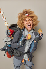 Emotional curly haired female backpacker spends free time at adventure camp holds cookies exclaims loudly keeps eyes closed carries backpack with necessary things isolated over grey background