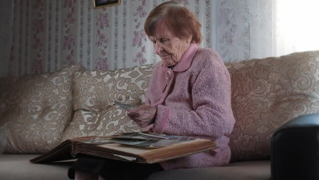 Elderly woman looks at photos in a family photo album. Old grandmother holds and looks at photographs with nostalgia, sitting on the sofa in the living room. Memories, biography or history concept