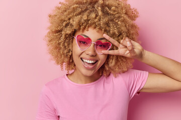 Portrait of happy curly haired woman makes peace gesture over eye smiles toothily has glad...