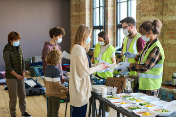 Group of young volunteers in masks and gloves spreading cooked food among refugees standing in front by table with containers