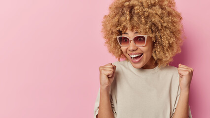 Positive surprised woman with curly hair celebrates victory roots for someone wears sunglasses and t shirt isolated over pink background feels motivated becomes champion triumphs over something