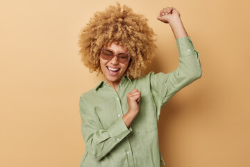 Cheerful curly haired woman shakes arms dances carefree has fun feels energetic dressed in casual...