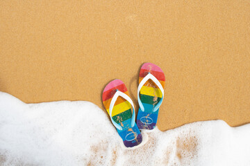LGBTQ flag flip-flop in a sand and a ocean wave. Top view of a wave in a sandals