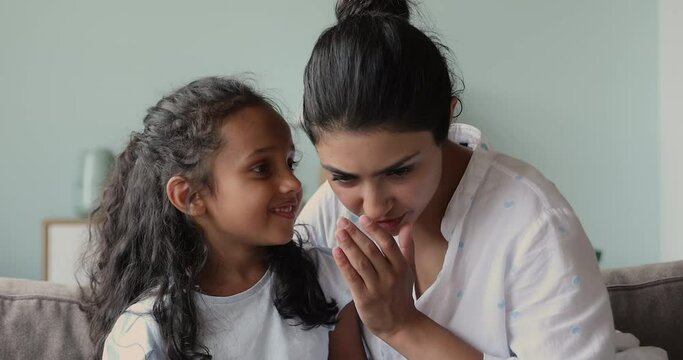 Happy two generations Indian family mom small girl relax on couch share secrets whisper funny rumors on ear laugh on hilarious tales. Grown elder small younger sisters enjoy telling confidential news