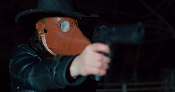 Criminal woman in plague doctor mask points gun to shoot at victim on dark background. Female killer holds dangerous weapon at night close view