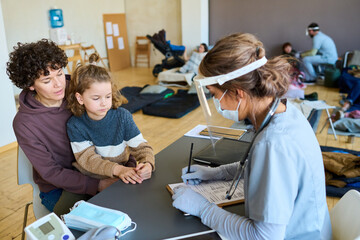 Young female clinician filling in medical form in front of woman with her little son sitting by...