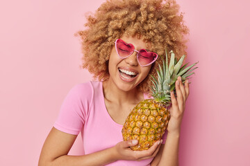 Overjoyed curly woman laughs out joyfully holds fresh pineapple going to make fresh juice from it poses with favorite exotic fruit dressed casually isolated on pink background. Healthy nutrition