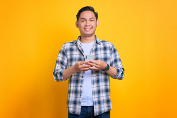 Smiling young Asian man in plaid shirt using mobile phone and looking at camera isolated on yellow background