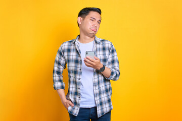 Disappointed young Asian man in plaid shirt using mobile phone, reacting to bad news isolated on yellow background