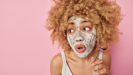 Close up shot of surprised young woman with blonde curly hair applies beauty mask holds eyelashes curler looks amazed away keeps mouth opened stands bare shoulders isolated over pink background