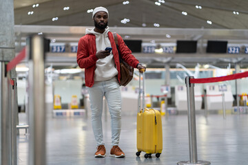 African American passenger man waiting to check in and drop off luggage in airport terminal, checks time of flight online, stands with suitcase, using mobile phone. Transportation, traveling concept.  © DimaBerlin