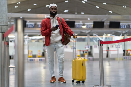 African American passenger man waiting to check in and drop off luggage in airport terminal, checks time of flight online, stands with suitcase, using mobile phone. Transportation, traveling concept.