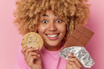 Happy curly haired European woman holds chocolate bar and cookie enjoys sweet life smiles happily...