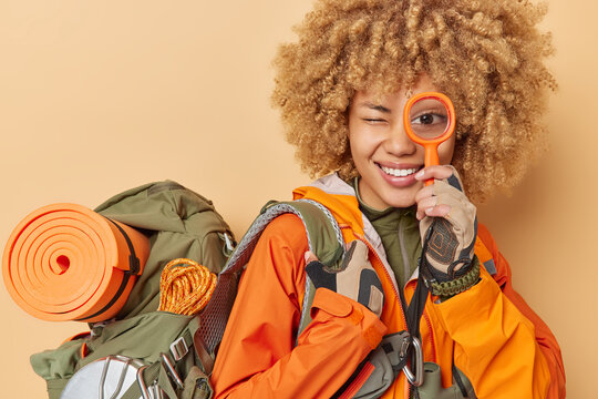 Positive curly female tourist smiles happily holds magnifying glass over eye explores new place carries backpack with necessary items for travel isolated over brown background. Woman backpacker