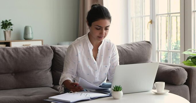 Millennial woman of Indian ethnicity work with documents from home analyze information using laptop pc take brief notes to daily planner. Busy young lady read business papers prepare financial report