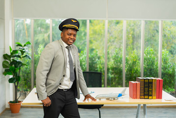Smart african businessman stands, wears pilot hat, looks at camera with airplane model on table and...