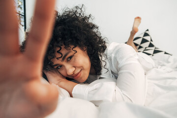 Shy young mixed race woman looking at camera lying down on the bed covering lens with hand....