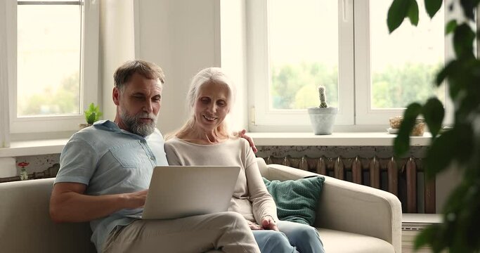 Older age couple on pension use laptop computer app sitting on sofa at living room. Mature husband confident pc user make internet purchases from home order consumer goods together with elderly wife