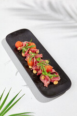 Roast beef with cherry tomatoes, ruccola and spicy sauce on black plate. Sliced roast beef on white concrete background. Italian antipasti in summer style with hard shadow and palm leaves.