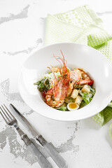 Classic Caesar salad with shrimps in white plate on stone table. Italian menu in restaurant. Seafood caesar on rustic style. Trendy salad with grilled prawns, green leaves, eggs, croutons and cheese.