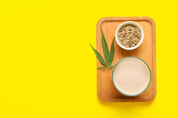 Wooden board with glass of hemp milk and seeds on yellow background