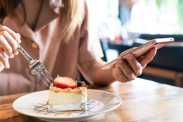 Asian woman using mobile phone while eating cake on wooden table in cafe. Relaxation, technology and lifestyle concept. Close up