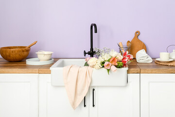 Sink with beautiful flowers and kitchen utensils on counters near lilac wall