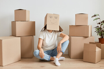 Full length portrait of Caucasian woman wearing funny cardboard box on head sitting on floor at new apartment, having fun alone while relocating to a new house.