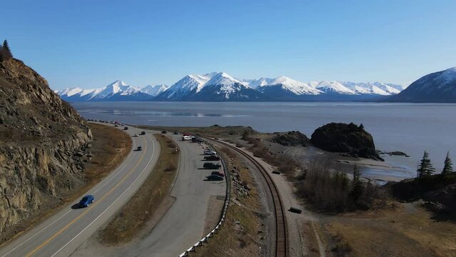 4k drone ascending above a parking lot and railroad tracks by the shoreline showcasing white capped mountains on a sunny day with a view of the bay in anchorage Alaska