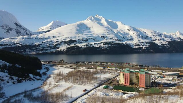 4K cinematic drone view over the town of Whittier Alaska showing a tall rise apartment building with  large whitecapped mountains in the background during winter