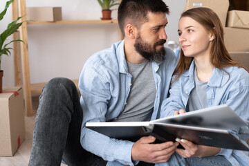 happy young couple on moving day sitting on floor in casual clothes hugging looking at photo album,...