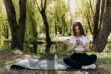 Fit pretty young woman chatting, browsing social media online on phone while sitting on yoga mat on green grass in park. Smiling caucasian sportswoman using smartphone outdoors.