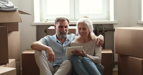 Excited elderly couple sit on floor among boxes with stuff at moving day use pad choose interior design for new flat. Happy aged spouses become homeowners rest from unpacking belongings with tablet pc - Powered by Adobe