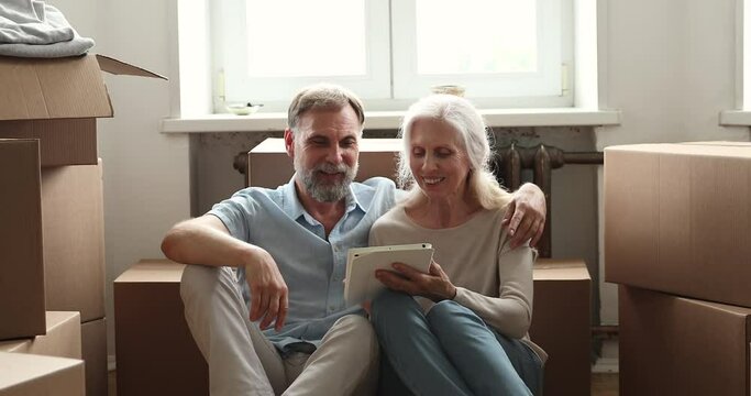 Excited elderly couple sit on floor among boxes with stuff at moving day use pad choose interior design for new flat. Happy aged spouses become homeowners rest from unpacking belongings with tablet pc
