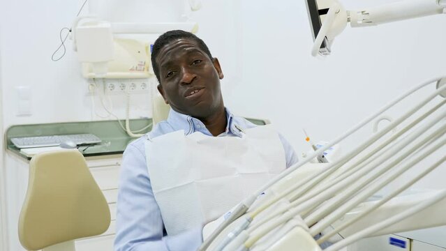 Portrait of positive african-american man sitting on dental chair and pointing at his teeth.