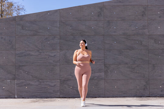 Smiling plus size woman listening music through wireless headphones walking in front of wall