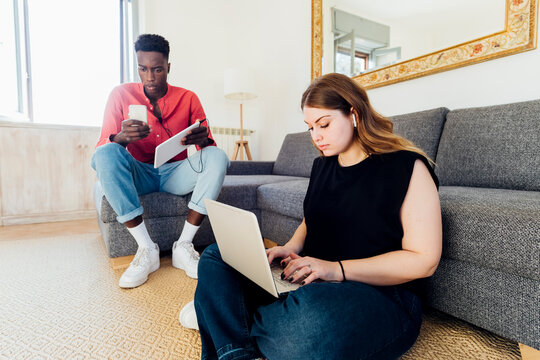 Multiracial couple using laptop and mobile phone in living room at home