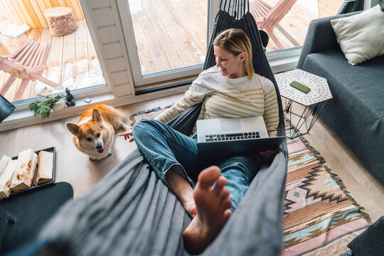 Woman playing with dog relaxing on hammock with laptop at home