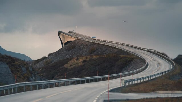 Storseisundet bridge on the Atlantic road rises above the landscape. Heavy clouds in the sky. Seagulls fly low. Slow-motion, pan left.