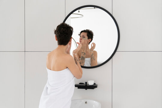 Woman wrapped in towel applying eye mask looking at mirror