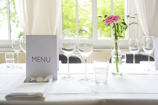 Elegant table setting, place with menu card, glasses, cutlery, napkin and a pink flower on a white tablecloth for a festive dinner celebration, copy space, selected focus