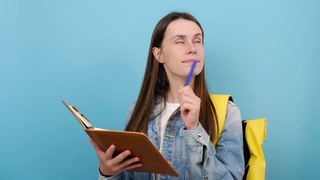 Portrait of pensive girl teen student wears denim jacket and yellow backpack writing down in exercise book notes, isolated on blue color background wall in studio. Education in university college