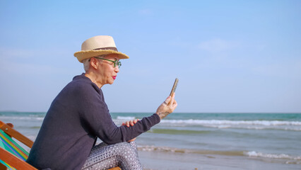 Asian elderly woman chatting with friends on video call while relaxing at the beach
