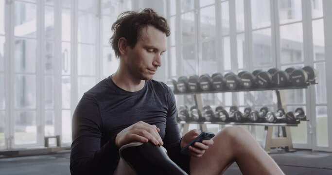 Caucasian male with prosthetic leg texting on his cellphone. Disabled athlete working out in modern-style gym.