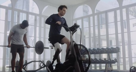 Caucasian male with prosthetic leg cycling on air bike while biracial male lifts weights in background. Disabled athlete working out in modern-style gym. - Powered by Adobe