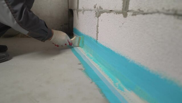 A worker is applying waterproofing paint to the floor in the bathroom. The master with a brush applies a protective layer of waterproofing to the floor.